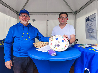 Links Uwe Klüter in einem blauen Pullover mit gelben Europasternen und einer blauen Kappe. Rechts Alexander Börger in einem weißen T-Shirt. Vor ihnen auf einem mit einer blauen Tischdecke bedeckten Tisch ein Glücksrad mit Flaggen europäischer Länder und ein Korb. Rechts von ihnen ein weiterer Tisch mit Informaterial zu Europa, der EU und der EUD. 