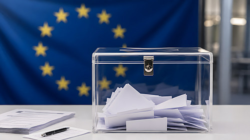 Eine transparente, mit Wahlzetteln etwa halb gefüllte Wahlurne auf einem weißen Tisch vor einer nicht fokussierten Europaflagge.